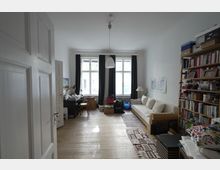 Wohnzimmer mit Holzboden, einer großen Fensterfront mit dunklen Vorhängen, einem Sofa an der rechten Wand, einem Tisch mit Stuhl auf der linken Seite sowie einem Bücherregal. Im Raum befinden sich auch Kartons und andere Gegenstände auf dem Boden.