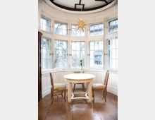 Ein rundes Zimmer mit großen, halbkreisförmig angeordneten Sprossenfenstern, die viel Tageslicht hereinlassen. In der Mitte steht ein weißer runder Tisch mit drei unterschiedlichen Stühlen, über dem eine dekorative sternförmige Hängelampe hängt; der Boden besteht aus Holzparkett.