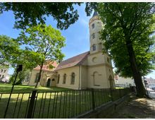 Gelbe Kirche mit rotem Ziegeldach und achteckigem Turm, umgeben von hohen Bäumen und einer Rasenfläche. Im Vordergrund steht ein schwarzer Metallzaun, der das Gelände abgrenzt.