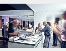 Moderne Küche mit einer großen Arbeitsinsel in heller Umgebung, in der mehrere Personen mit Schürzen zusammen kochen. Im Vordergrund stehen Küchenutensilien, Schüsseln und Zutaten, während der Hintergrund durch Arbeitsflächen und Küchenschränke geprägt ist.