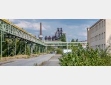 Industriegelände mit langen grünen Rohrleitungen auf Metallstützen, einem gepflasterten Weg im Vordergrund und Industrieanlagen mit Schornstein und Hochöfen im Hintergrund. Rechts steht ein beige-farbenes Verwaltungsgebäude, und das Gelände ist von Büschen und vereinzelten Bäumen umgeben.