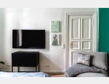 Wohnzimmer mit einer weißen Wand, einem Flachbildfernseher, einem schwarzen, niedrigen Schrank und dekorativen Bildern. Rechts eine weiße Tür mit Kassettenmustern, daneben ein graues Sofa mit einem Kissen und einer Decke, auf dem Boden heller Holzfußboden.