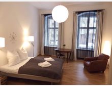 Schlafzimmer mit einem Doppelbett, dunkler Tagesdecke und weißen Kissen, daneben zwei Stehlampen. Im Hintergrund sind zwei große Fenster mit Vorhängen, ein runder Holztisch mit einem Stuhl und ein brauner Ledersessel. Der Raum hat Holzboden und eine moderne Einrichtung.