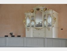 Weiße Kirchenorgel mit goldenen Verzierungen und glänzenden Pfeifen, eingebaut in eine schlichte, helle Kirchenwand über einer mit weißem Holz vertäfelten Empore.