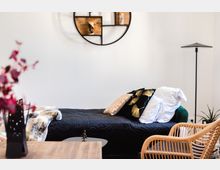 Ein modernes Wohnzimmer mit einem Tagesbett mit schwarzer gesteppter Decke und dekorativen Kissen in Schwarz und Gold. Im Vordergrund steht ein Holztisch mit einer schwarzen Vase und roten Zweigen, im Hintergrund eine runde, minimalistische Wandregalstruktur und eine Stehlampe.