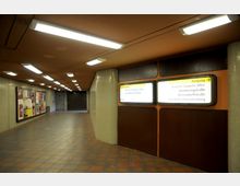 U-Bahn-Unterführung mit orange-braunen Wänden und Decken, beleuchtet durch rechteckige Deckenleuchten. An den Wänden befinden sich beleuchtete Wegweiser mit Zielangaben sowie eine Fläche mit Plakaten.
