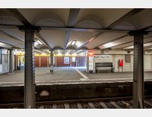 U-Bahnhof mit offener Bahnsteighalle im Station Theodor-Heuss-Platz, gekennzeichnet durch wellenförmige Decken, tragende Metallpfeiler und ein beleuchtetes SOS-Zeichen. Im Hintergrund sind geschlossene Türen, Informationenstafeln, eine Bank und ein Feuerlöscher sichtbar.
