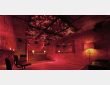 Ein kleiner Clubraum mit Holzwänden, einer erhöhten DJ-Konsole im Hintergrund und einer gemütlichen Beleuchtung in warmem Rot. Die Decke ist mit leuchtenden LED-Lichtern verziert, während auf der rechten Seite ein großer Spiegel und eine Stehlampe sichtbar sind.