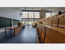 Ein moderner Hörsaal mit ansteigenden Holzsitzreihen, einer großen Fensterfront, die Tageslicht hereinlässt, und einer Schultafel sowie einem Rednerpult im vorderen Bereich. Im Fenster sind Außenansichten eines Backsteingebäudes sichtbar.