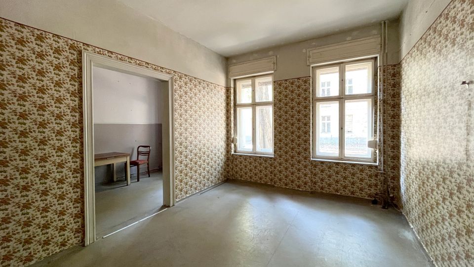Leerer Innenraum in einem älteren Gebäude mit abgenutztem Boden und floral gemusterter Tapete an den Wänden. Zwei große Sprossenfenster lassen Tageslicht herein; links führt ein breiter Durchgang in einen Nebenraum.