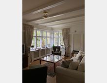 Wohnzimmer mit großen Sprossenfenstern, durch die Tageslicht einfällt. Zur Einrichtung gehören ein beigefarbener Sofa, ein Glastisch, ein Sessel, Vorhänge, ein Teppich und Dekorationsgegenstände auf einer Fensterbank.