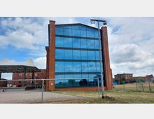 Ein modernes Gebäude aus rotem Backstein mit einer Glasfassade, die den Himmel reflektiert. Im Vordergrund befindet sich eine asphaltierte Fläche, daneben ein Metallzaun, und im Hintergrund sind weitere Backsteingebäude sowie eine wieseartige Fläche erkennbar.