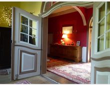 Ein Blick in einen Innenraum durch eine geöffnete Doppeltür mit geschwungenen Holzverzierungen, dahinter ein Raum mit roter Wand, Holzanrichte, Tischlampe und dekorativem Teppich. Ein gebogener Türrahmen ergänzt die klassische Einrichtung.