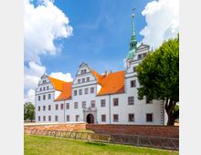 Weiße Schlossanlage mit markanten roten Ziegeldächern und zahlreichen Fenstern in symmetrischer Anordnung. Im Vordergrund befindet sich eine gepflasterte Mauer sowie eine Wiese mit einer Sitzbank, der Hintergrund zeigt blauen Himmel mit weißen Wolken.