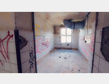 Verlassenes Zimmer mit gefliesten Wänden, bedeckt mit Graffiti in Rot und Gelb. Die Decke ist beschädigt, Putz fällt herab, ein großes Rohr ragt hervor, und ein Fenster mit geteilter Scheibe lässt Tageslicht herein.