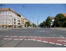 Straßenkreuzung in einer Stadt mit Straßenbahnschienen, flankiert von mehrstöckigen Wohngebäuden mit Geschäften im Erdgeschoss links und Bäumen sowie Gehwegen rechts. Oberleitungen für Straßenbahnen und eine klare, strukturierte Fahrbahn sind sichtbar.