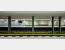 U-Bahn-Station mit zwei Bahnsteigen, getrennt durch Gleise und grünen Säulen. Die Wände sind hellgrün gestrichen, mit Sitzbänken, einem Stationsschild ("Kurfürstendamm") und Plakaten an den Seiten.
