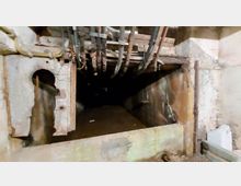 Ein verfallener, unterirdischer Gang mit rostigen Metallrahmen, freiliegenden Rohren an der Decke und bröckelnden Betonwänden. Der Bereich wirkt dunkel und verlassen, mit sichtbaren Verfallsspuren.