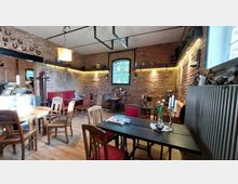 Ein gemütliches Café mit Backsteinwänden, Holztischen und einer Kombination aus modernen und antiken Möbeln. Im Raum stehen dekorative Elemente wie Kerzenständer, Tulpenvasen und ein Kronleuchter, während durch ein Gitterfenster Tageslicht einfällt.