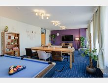 Wohnraum mit gemütlicher Einrichtung, bestehend aus einem Holzesstisch mit gemischten Stühlen, einem Sofa, einem Fernseher an einer lila Wand und dekorativen Pflanzen. Im Vordergrund ein Billardtisch, und rechts große Fenster mit weißen Vorhängen, die viel Tageslicht hereinlassen.