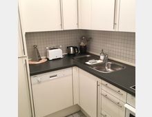 Eckküche mit weißen Schränken und dunkler Arbeitsplatte, grauer Fliesenrückwand und Edelstahlspüle mit Abtropffläche. Auf der Arbeitsfläche stehen Küchenutensilien wie Toaster, Wasserkocher und Reibe; unten sind Geschirrspüler und Schubladen integriert.