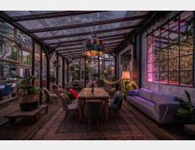 Ein gemütlicher Wintergarten mit hölzernem Boden und einer teilweisen Glasdecke, ausgestattet mit einem langen Holztisch, umgeben von unterschiedlichen Stühlen, einer Couch und Pflanzen. Die Wände bestehen aus Glas und Ziegeln, und eine bunte Hängelampe sowie dekorative Beleuchtung sorgen für stimmungsvolle Atmosphäre.