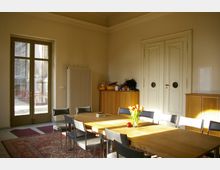 Heller Raum mit einem großen Holztisch und mehreren Stühlen, ein Strauß Tulpen und ein Apfel stehen auf dem Tisch. Im Hintergrund sind eine hohe Doppeltür, ein Heizkörper, ein Schrank sowie eine verglaste Tür mit Blick auf einen Außenbereich zu sehen.