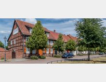 Fachwerkhäuser mit roten Ziegeldächern an einer ruhigen, von Bäumen gesäumten Straße. Im Vordergrund sind gepflasterte Gehwege, parkende Autos und eine Laterne zu sehen.