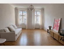 Bright, minimalist living room with light wood floors, a white sofa along one wall, and a low wooden media console on the opposite side. Two tall windows with sheer curtains and radiators beneath them face the street, with a glass globe pendant light hanging from the ceiling.