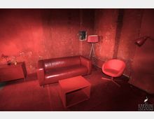 Roter Raum mit Sichtziegelwänden und einer Einrichtung aus roten Möbeln, darunter ein Ledersofa, ein Sessel, ein Beistelltisch und eine Stehlampe. Links steht ein niedriger Schrank mit Dekoration, auf dem Teppichboden.