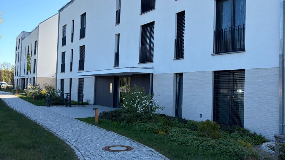 Moderner, weißer Mehrfamilienhauskomplex mit mehreren Stockwerken, großen rechteckigen Fenstern und teils schwarzen Balkongeländern. Ein geschwungener gepflasterter Gehweg führt durch begrünte Vorgärten zum überdachten Hauseingang.