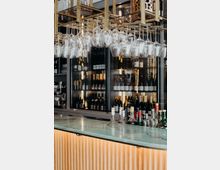 Eine moderne Bar mit einer beleuchteten Theke aus Holz und einer gläsernen Oberfläche. Im Hintergrund sind ein Weinkühlschrank mit Flaschen und ein Regal mit hängenden Weingläsern sichtbar.