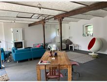 Großer, offener Wohnraum mit industriellen Elementen wie freiliegenden Stahlträgern und einer weiß verputzten Decke. Der Raum enthält eine Sitzecke mit blauen Sofas, einen modernen Kamin, einen hölzernen Esstisch, ein Bett in einer Ecke und einen auffälligen weißen Schalensessel mit roter Polsterung.