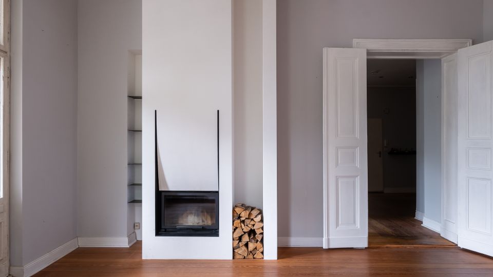 Heller, leerer Wohnraum mit hohen, hellgrauen Wänden und Holzdielenboden, in der Mitte ein weißer Kamin mit gläsernem Einsatz und daneben gestapeltem Brennholz. Rechts führen geöffnete, weiße Doppeltüren in einen angrenzenden Raum; links sind schmale, eingebaute Regale zu sehen.