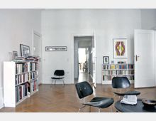 Wohnzimmer mit weißen Wänden, Holzdielenboden und minimalistischer Einrichtung. Bücherregale stehen an den Wänden, zwei schwarze Design-Stühle sowie ein runder Couchtisch befinden sich im Raum, und durch eine geöffnete Tür ist ein Flur sichtbar.