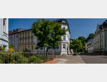 Straßenszene in einem Wohnviertel mit historischen, mehrstöckigen Stadthäusern im Stil der Gründerzeit, hell gestrichenen Fassaden und Sprossenfenstern; im Vordergrund ein Garten mit schmiedeeisernem Zaun und ein großer Baum. Die gepflasterte Straße ist ruhig und wird von weiteren Bäumen und Laternen gesäumt.