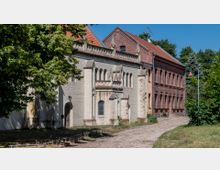 Ziegelsteingebäude im historischen Stil mit verzierten Spitzbögen, daneben ein weiteres Gebäude aus rotem Backstein. Eine gepflasterte Straße führt entlang der Häuser, umgeben von Bäumen und Grünflächen.