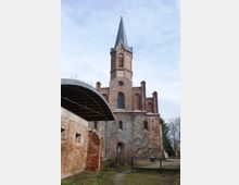 Backstein- und Feldsteinkirche mit einem hohen, schlanken Turm und spitzem Dach, im Vordergrund eine gemauerte, teils verfallene Wand und ein kleiner Weg, der zu einem Eingangstor führt. Der Himmel ist leicht bewölkt, und Bäume umgeben die Szene.