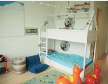 Ein Kinderzimmer mit einem weißen Hochbett in Hausform, das mit blauer Bettwäsche und Mondmotiven dekoriert ist. Im Raum befinden sich auch bunte Spielzeugfiguren, ein Sitzsack, ein geflochtener Korb, ein Teppich mit Straßenmotiv und Wandbilder, umgeben von hellem Holzboden.