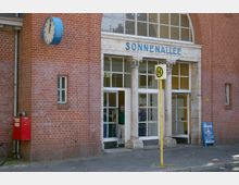 Backsteinfassade eines Gebäudes mit der Beschriftung „SONNENALLEE“ über dem Eingang, flankiert von dekorativen Säulen. Links ein blaues Uhrengehäuse und ein roter Briefkasten, rechts ein gelbes Haltestellenschild mit grünem „H“.