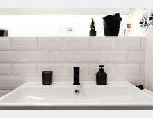 Weißes Badezimmerwaschbecken mit schwarzen Seifen- und Dekorflaschen darauf, vor einer Wand mit glänzenden weißen Fliesen im U-Bahn-Stil; obenauf ist eine Ablage mit kleinem Pflanzengefäß, geflochtenem Korb und diffuser Dekoration.