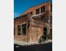 Red-brick industrial-style building with tall multi-pane windows and weathered masonry, set along a paved courtyard. A glass entryway with potted plants sits at the corner beneath a sloped roofline and upper-level windows.