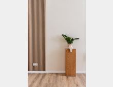 Innenraum mit heller Wand, einer Holzlattenverkleidung auf der linken Seite und einem Holzboden. Rechts steht ein schmaler Holzsockel, auf dem ein weißer Blumenkübel in Form eines sitzenden Menschen mit grüner Pflanze platziert ist.