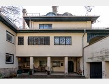 Hinterhof einer zweistöckigen, historischen Villa mit hellem Putz, Gitterfenstern und einer überdachten Terrasse, die von drei Säulen gestützt wird. Im Erdgeschoss befinden sich Sitzgelegenheiten und Pflanzen, seitlich schließt eine Garage an.