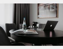 Ein moderner Besprechungsraum mit einem runden, schwarzen Holztisch, umgeben von gepolsterten Stühlen. Auf dem Tisch liegen ein offenes Buch, eine Glasflasche Wasser mit einem Glas, ein aufgeklappter Laptop und Schreibutensilien, im Hintergrund ein Vorhang und eine Kaffeemaschine.