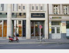 Fassade eines Gebäudes in der Oranienstraße 164 mit großen Fenstern und einer Eingangstür im Erdgeschoss, daneben ein Schild für ein Yoga-Studio und ein geparktes Fahrrad sowie ein Motorroller auf dem Gehweg. Ein Parkverbotsschild steht vor dem Gebäude.