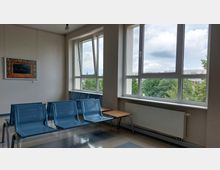Wartebereich mit blauen Plastikstühlen und kleinen Holztischen, großer Fensterfront mit Blick auf Bäume und Gebäudestrukturen im Hintergrund. An der cremefarbenen Wand hängt ein gerahmtes Kunstwerk, darunter ein Heizkörper.
