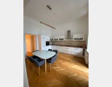 Moderne Küche mit weißer Einbauküche, hölzerner Arbeitsplatte und Dunstabzugshaube. In der Mitte des Raumes steht ein weißer Esstisch mit vier gepolsterten Stühlen auf einem Parkettboden im Fischgrätenmuster.