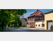 Straßenszene mit Kopfsteinpflaster, flankiert von grünen Bäumen links und einem roten Backsteingebäude mit rotem Ziegeldach rechts. Daneben ein gelbes Gebäude mit der Aufschrift "HF" und einem geschlossenen Rolltor. Im Hintergrund sind weitere Wohnhäuser und eine ruhige Umgebung erkennbar.