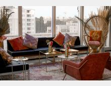 Wohnzimmer mit modernem, gemütlichem Design: Schwarzes Sofa mit farbigen Kissen, ein geflochtener Sessel mit dekorativem Kissen, sowie kleine goldene Beistelltische mit Blumenvasen und Kerzen. Im Hintergrund eine große Fensterfront mit Blick auf umliegende Gebäude und einen Balkon.